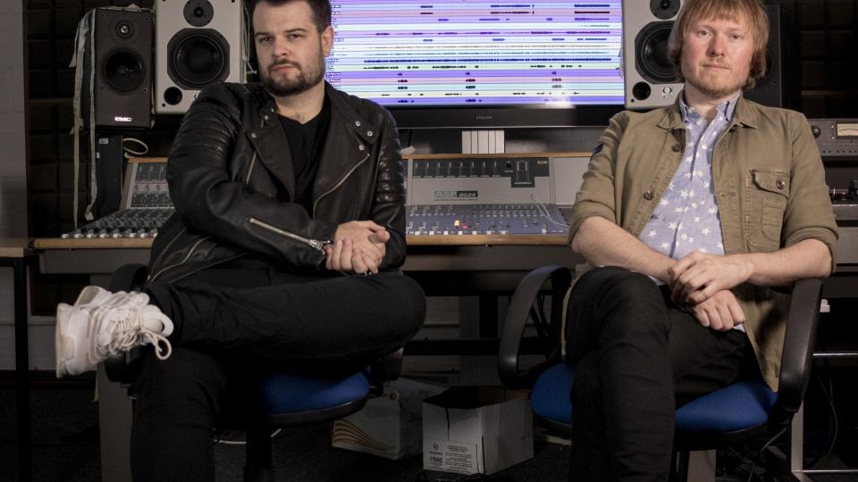 Dan and Ben sit in front of a mixing desk. Both have one leg crossed and are looking at the camera