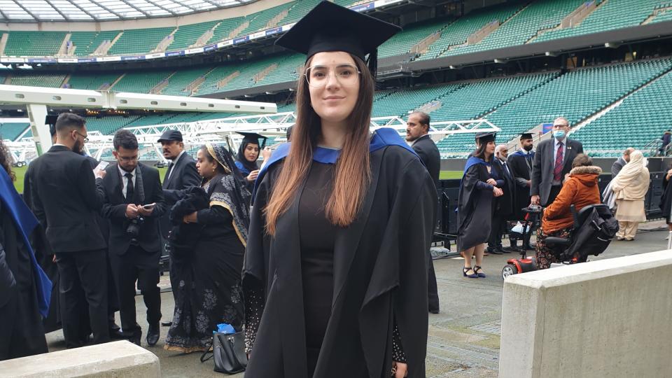 International student Andreea-Daniela Coroama standing in graduation clothes at her graduation ceremony in a sports stadium