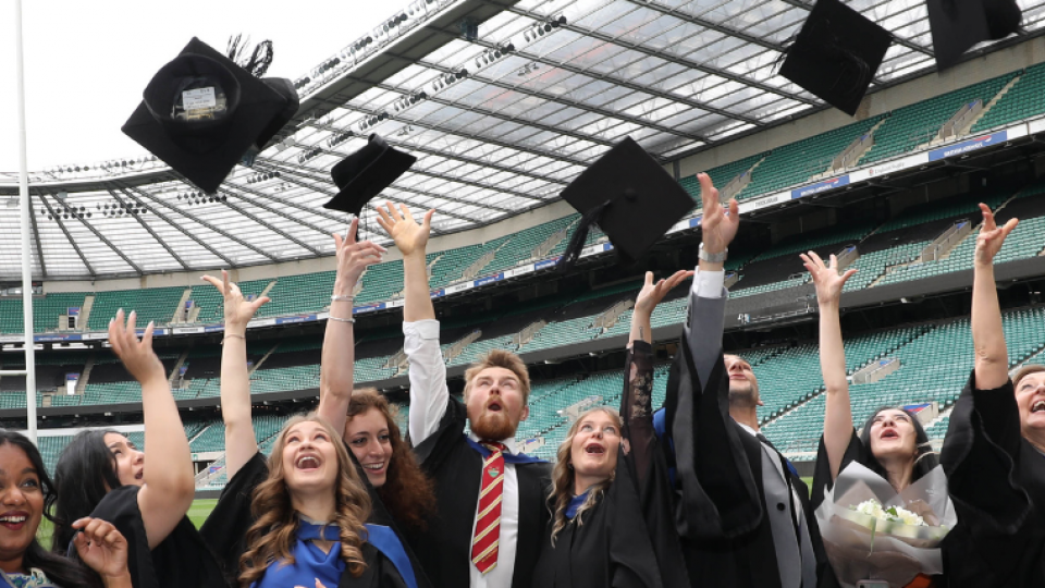 A group of graduating students are throwing their caps into the air. They are standing in front of the rugby pitch at Twickenham stadium. They are all wearing black gowns and formal dress.