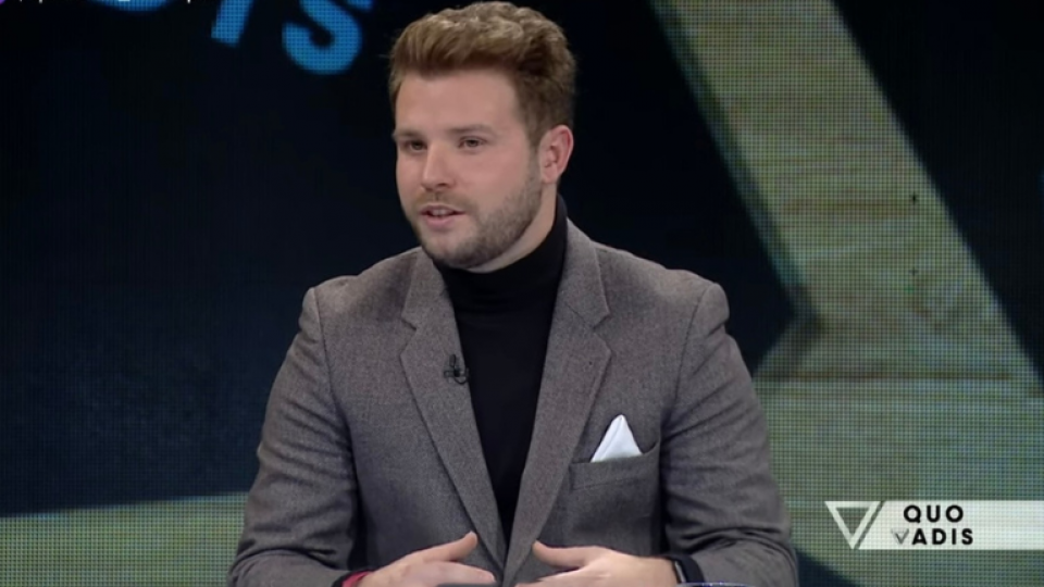 Daniel is wearing a grey suit with a black turtleneck underneath. He is stood in front of a large screen and is looking to the left. He has a microphone attached to his lapel and is giving a speech.