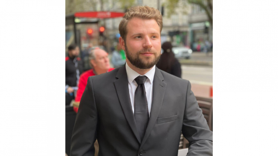 Daniel is wearing a dark grey suit with a white shirt and matching tie. He has light brown hair and beard. He is standing outside on a busy street. 