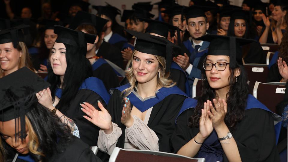 Graduands sat down and clapping during a ceremony, wearing gowns and mortarboard hats.