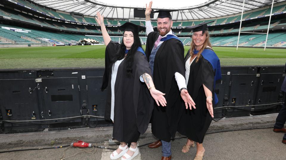 Three graduands posing with their arms out pitchside at Twickenham Stadium.