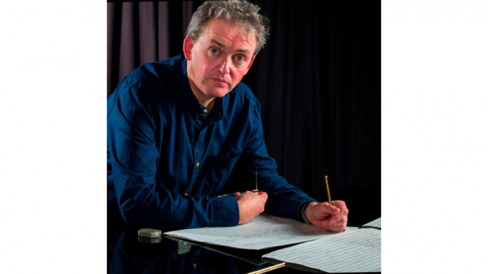 Francis is wearing a dark blue collared shirt and is leaning over a piano where a score is laid out in front of him. He is holding a pencil and is looking toward the camera.