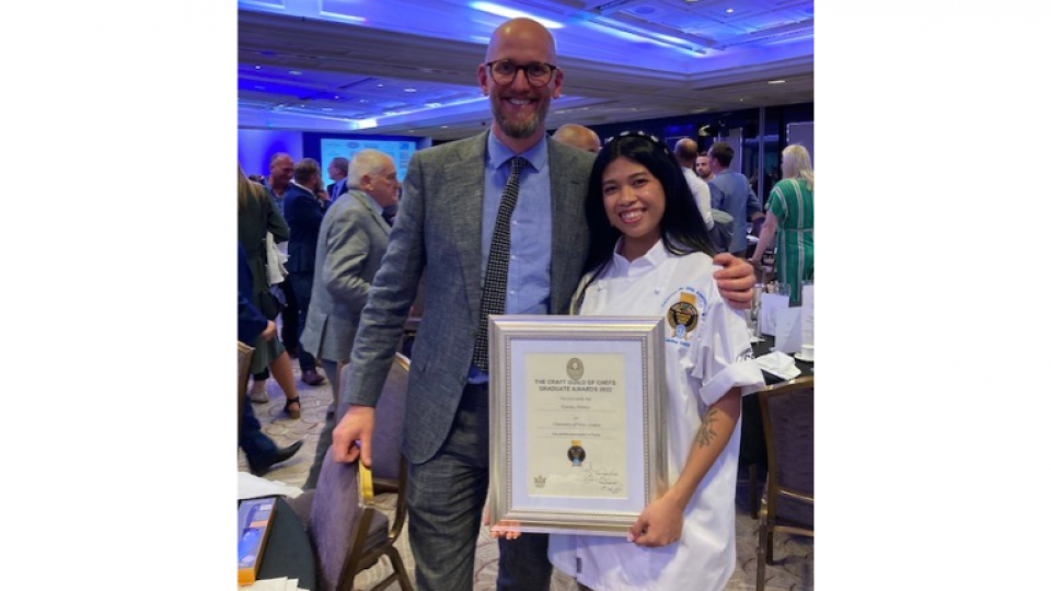 Katrina is wearing her chef overalls and holding a large framed certificate. Next to her stands Ben Christopherson her tutor wearing a grey suit.