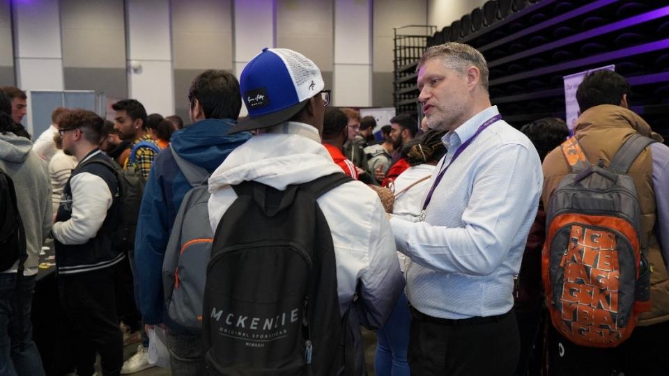 A student wearign a cap backwards is talking to someone from the careers fair wearing a shirt. The room is full of students and stalls.