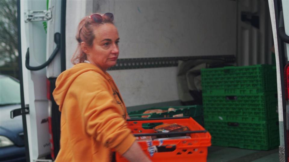 Rachel is wearing an orange hoodie and is unloading trays of food from the back of a van for her food bank charity.