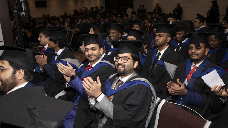 Students are sat in rows in a large room at Twickenham where the graduation ceremony is about to commence. All students are wearing the UWL cap and gown and are clapping.