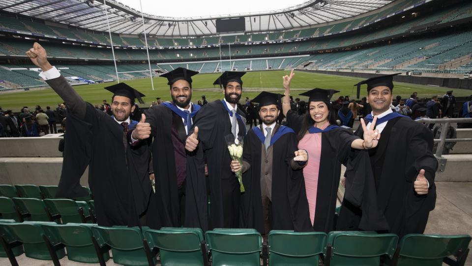 Group of 6 students wearing the UWL graduation cap and gowns are standing in the twickenham stands with the view of the stadium behind them. They are all smiling.