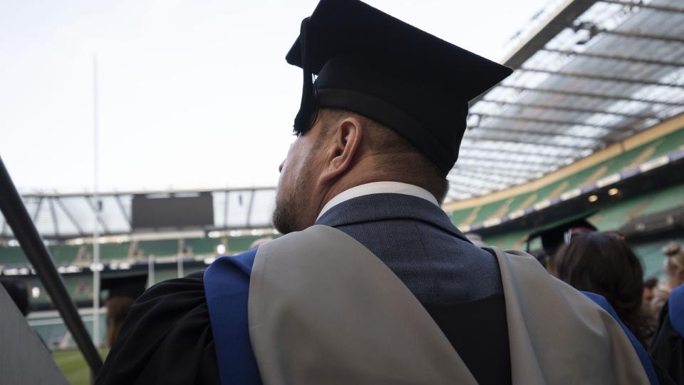 A student walking out towards the pitchside of Twickenham Stadium with his gown and hat on.