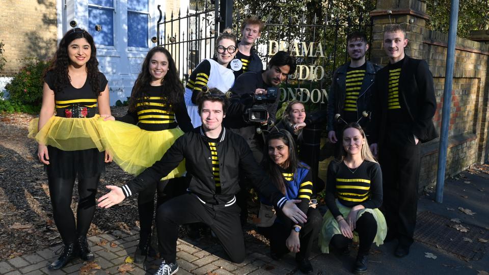 A group of Drama studio london students are dressed up in panto costumes ready to perform the bees knees. They are stood in front of the drama studio london gate and wearing black and yellow stripes with yellow tutus.