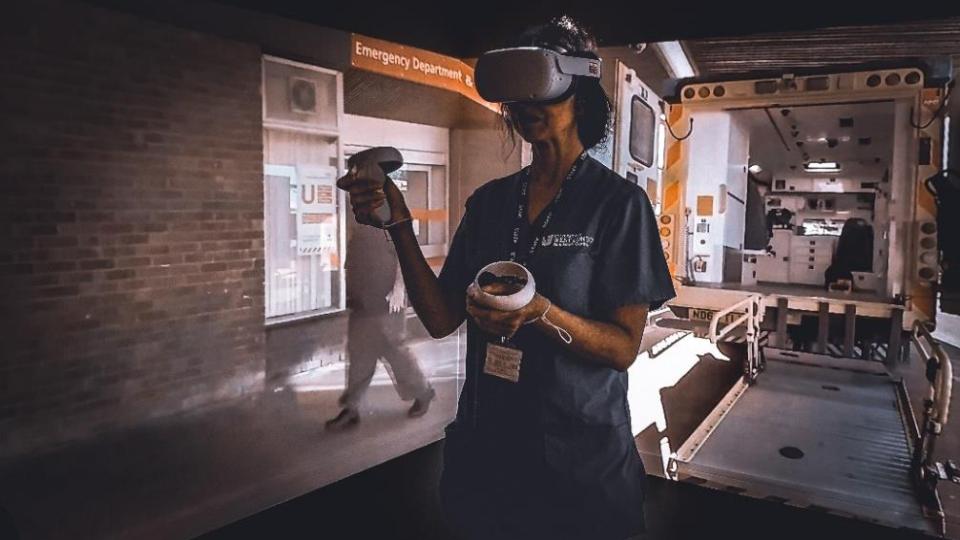 Medical professional in blue uniform wearing VR headset and holding controllers whilst standing in front of an ambulance..