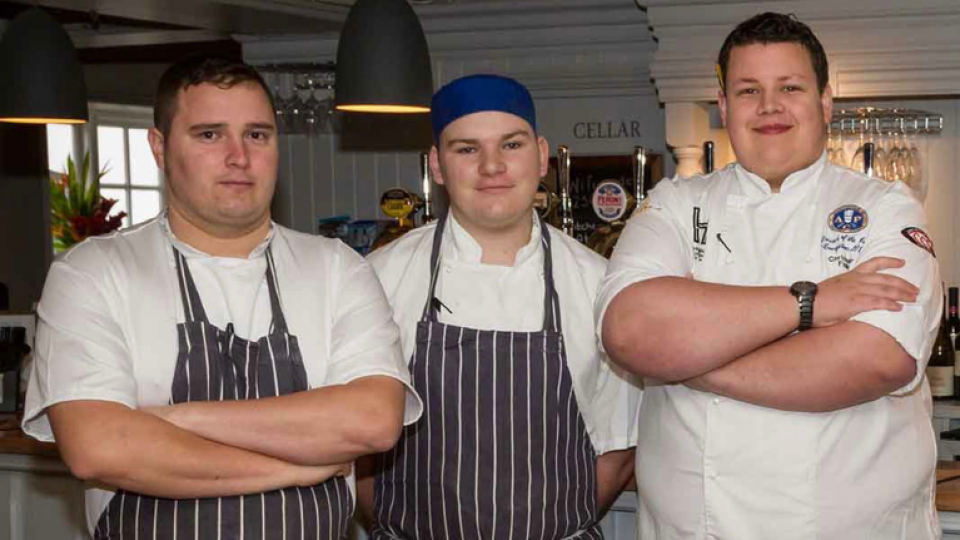 Chris Hurter, wearing white culinary clothing and crossing his arms, is standing with two other chefs in a restaurant.