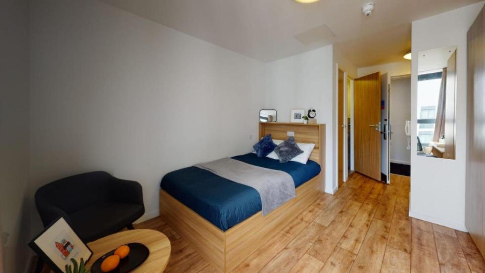 A modern room, with a double bed including blue and white sheets. There is laminate wooden flooring, a black chair and small table.