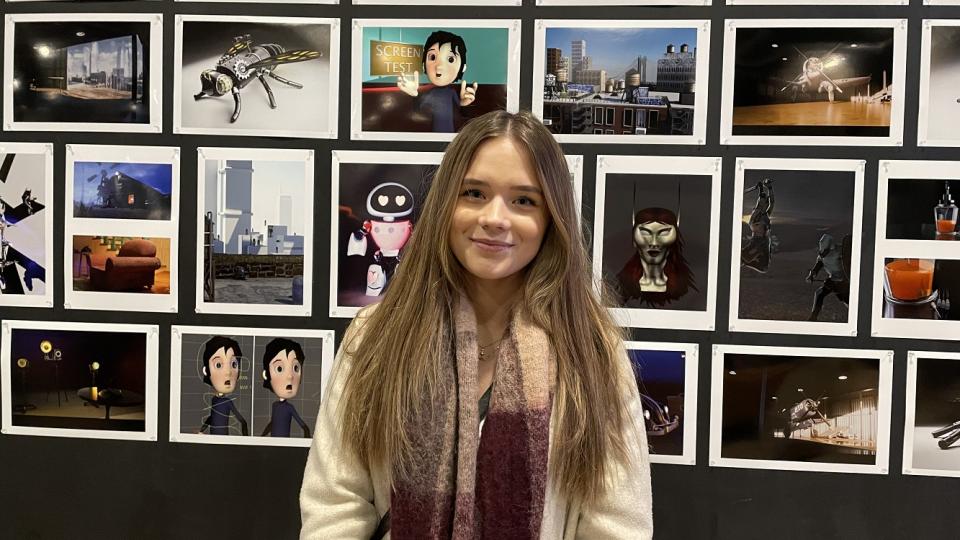 A student stands smiling in front of her Visual Effects work, printed and mounted on a black wall