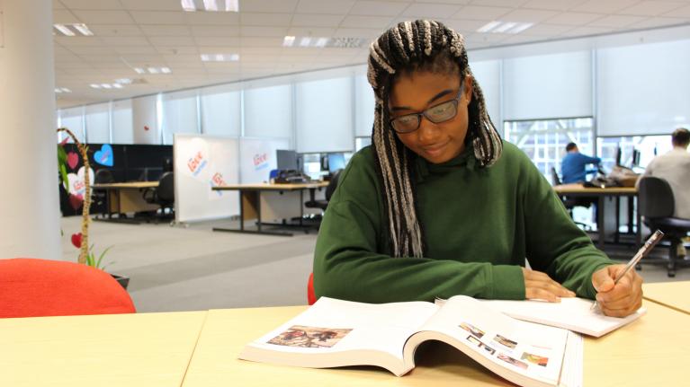 A female student wearing a green jumper and glasses is studying with books 
