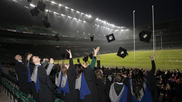 A group of graduates throwing hats into the air, pitchside at Twickenham Stadium, showing the grass and dark sky.