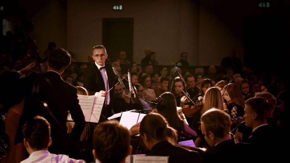 Composer Blazej Mokrzak standing amongst orchestra in front of seated audience