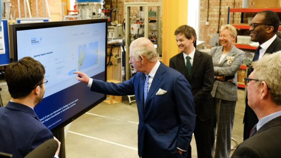 Prince Charles is wearing a navy blue suit and is standing looking and pointing at a television screen. He is surrounded by a group of people who are smiling and also looking at the screen. 