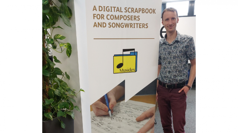Alex Liberatore is stood infront of a poster that says 'a digital scrapbook for composers and song writers'. Alex is wearing a patterned button up shirt and maroon trousers. 