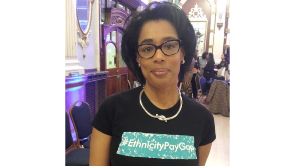 Sianne is wearing a top that says #ethnicitypaygap with a silver necklace. She is in a large room and there are chairs and tables set up behind her. She has shoulder length black hair and rectangle glasses.