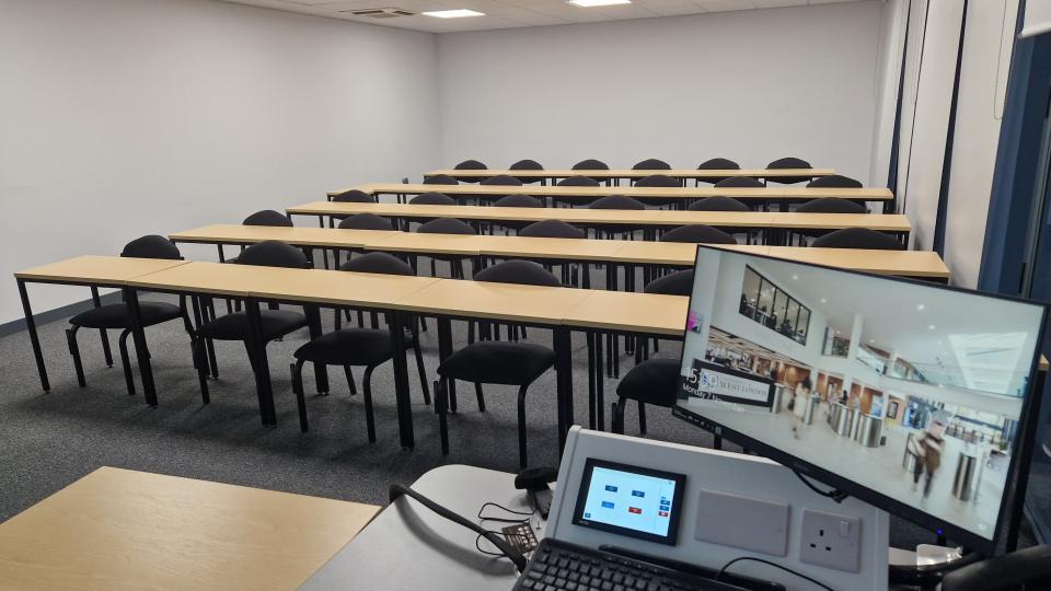 Rows of tables with black chairs behind are pictured in a large white room. There is a computer and desk at the front of the room.