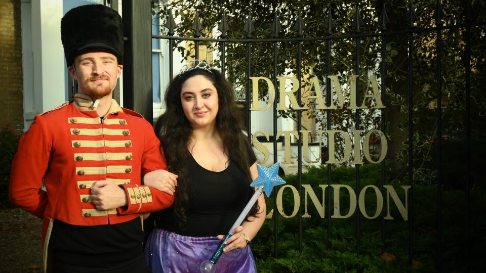 Two students in costume ready to perferm the nutcracker are stood outside the Drama studio London building. A man is wearing a red jacket and a small beefeater style hat. A woman has her arm linked through his and is holding a magic wand.