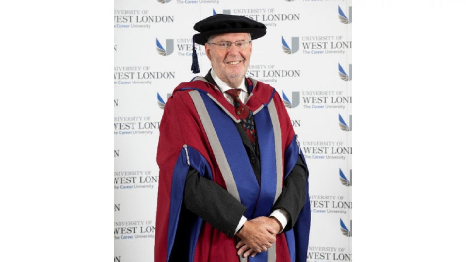 Martin is wearing the UWL official honorary robes, smiling at the camera infront of a wall with the UWL logo on.