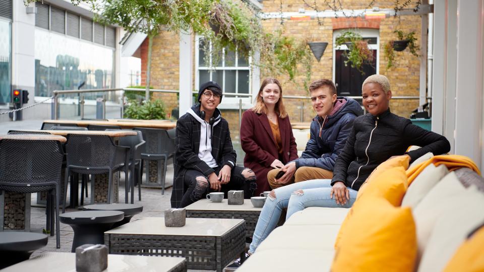 Four students seated outside with hot drinks, surrounded by hanging plants, tables and chairs.