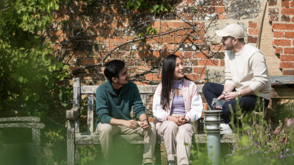 Two males students and one female student sit on a bench in a garden in the sun and are talking