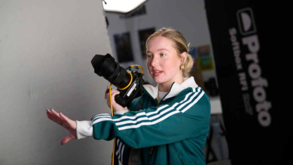A student directing people whilst holding an SLR camera, wearing a green sweatshirt.