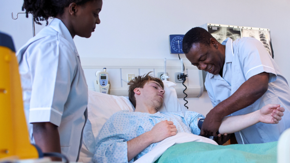 Male nurse tending to patient in hospital bed, with a second nurse present.