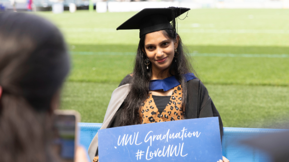 A graduate posing pitchside at Twickenham Stadium, holding a sign that says "UWL Graduation #LoveUWL"