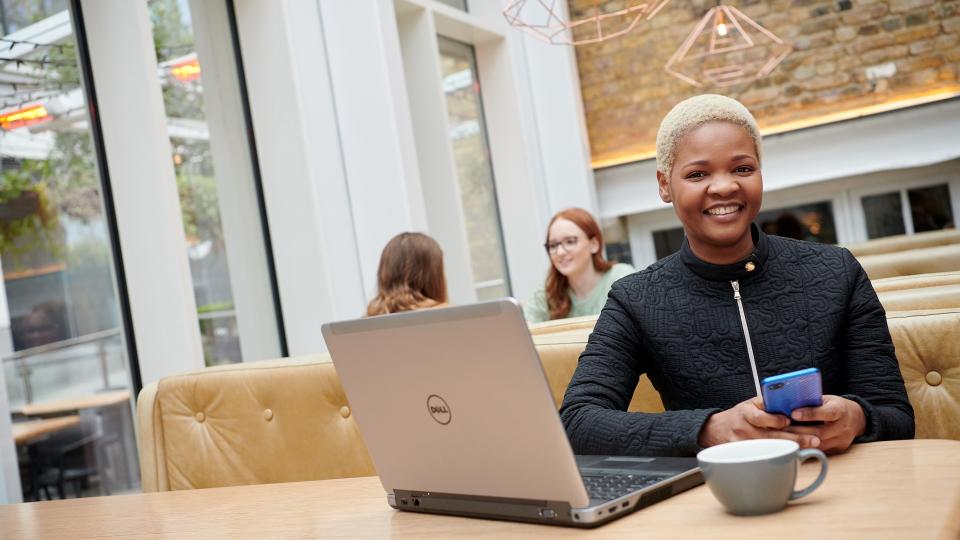 A student sitting in a cafe next to her laptop and holding her phone.