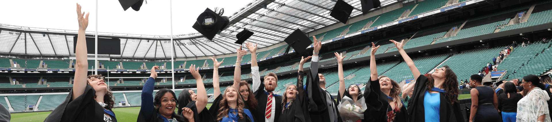 A group of graduating students are throwing their caps into the air. They are standing in front of the rugby pitch at Twickenham stadium. They are all wearing black gowns and formal dress.