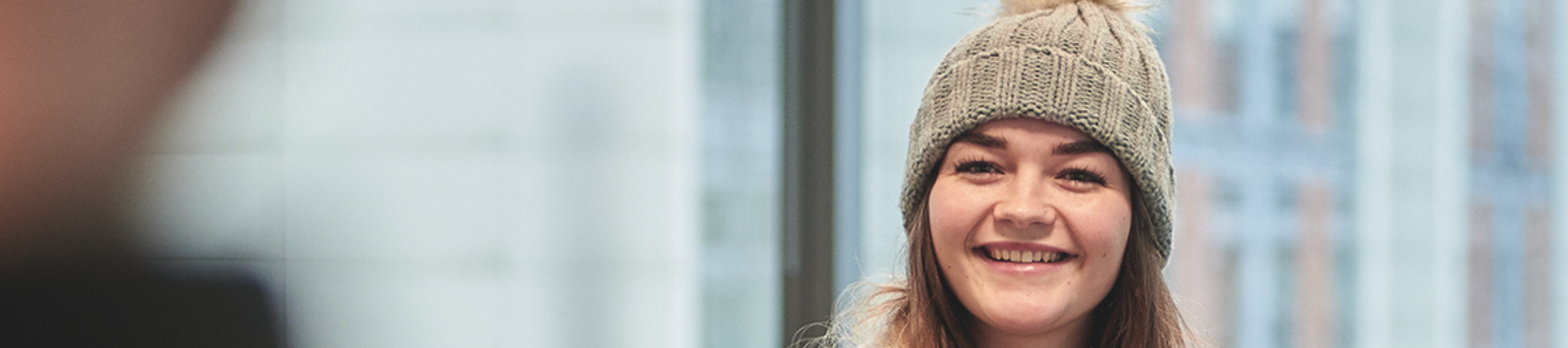 A student smiling, wearing a woolly bobble hat in front of a large window.