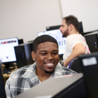 Students sat in a computer lab, one looking at his computer smiling.