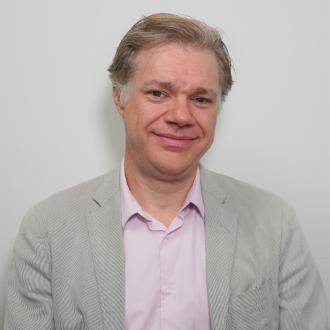 James is wearing a pale pink shirt and a grey suit. He has light brown and grey hair . He is smiling at the camera and has blue eyes.